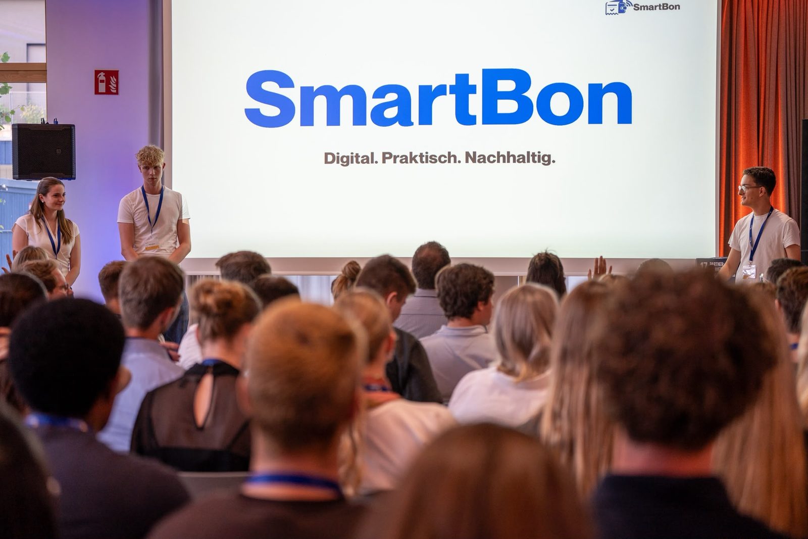 Das SmartBon-Team pitcht auf einer Bühne, auf der Leinwand steht SmartBon: Digital. Praktisch. Nachhaltig.